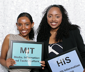 Two FIMS alumni holding signs that say MIT and HIS and 