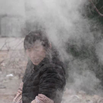 A person in a black coat standing outside, partly obscured by fog or steam.