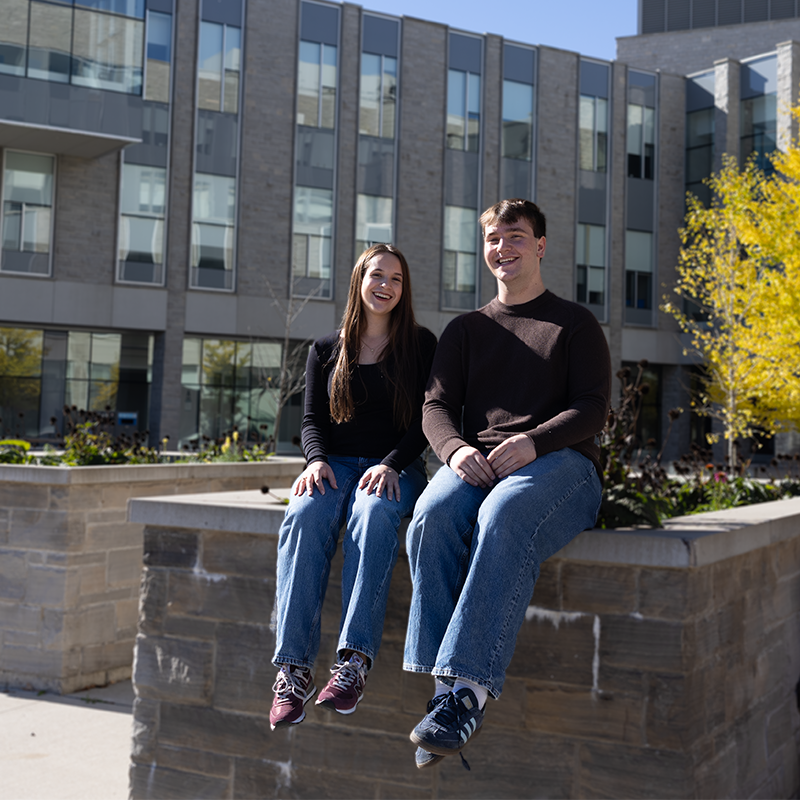 Two students sitting outside of the FIMS & Nursing Building on a sunny day