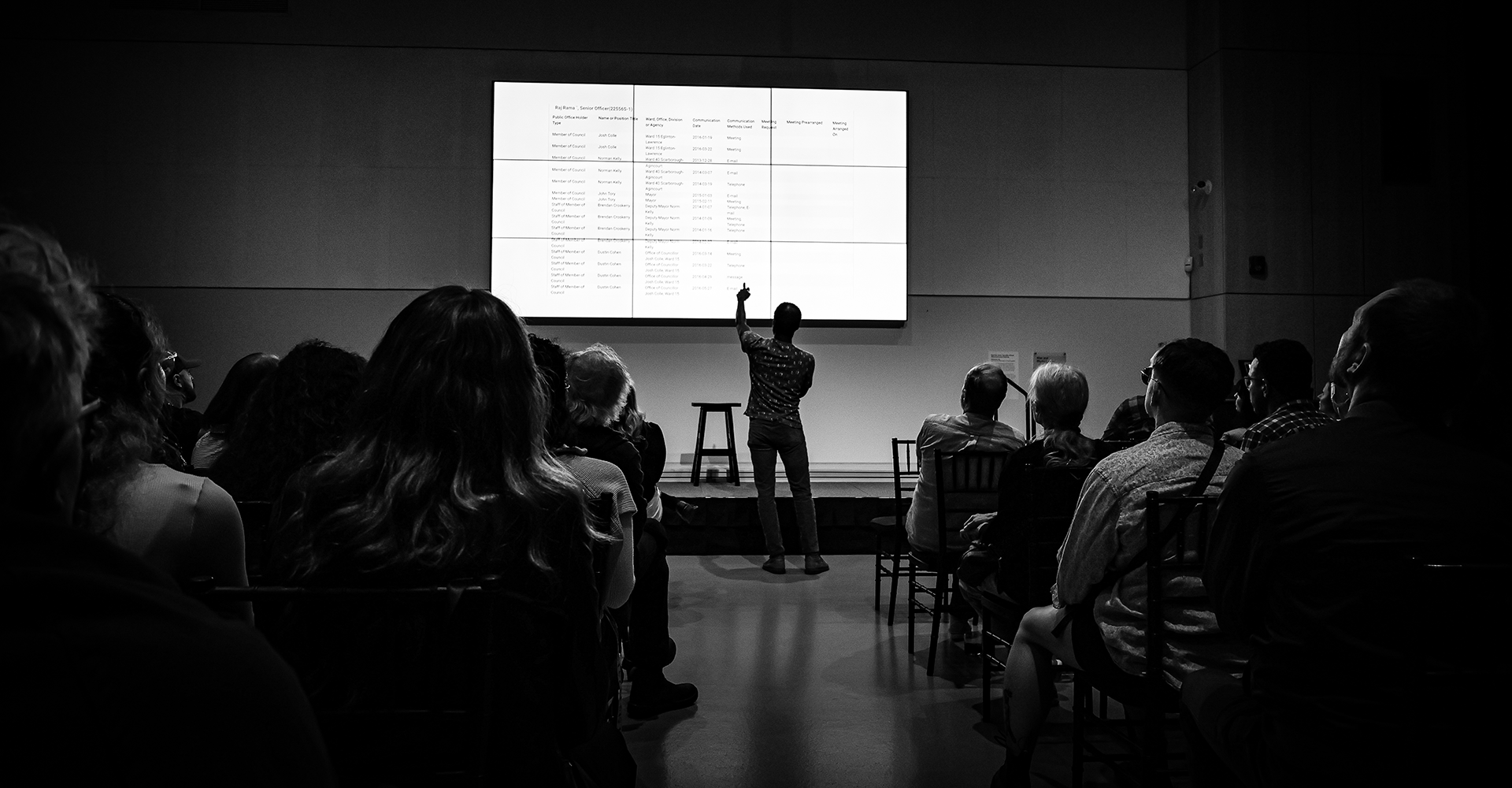 Black and white photo of a man lecturing a full room and pointing at the display screen
