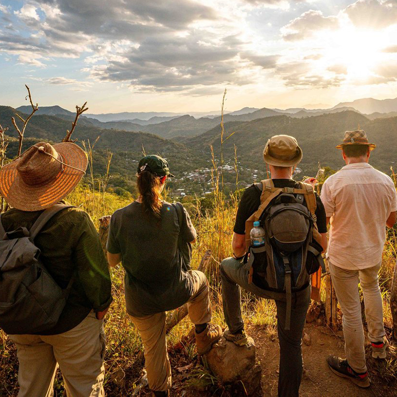 Several researchers with backpacks survey a small town from a hill top in El Salvador. The sun is setting. Photo by Shawn Robertson in 2023 in El Salvador