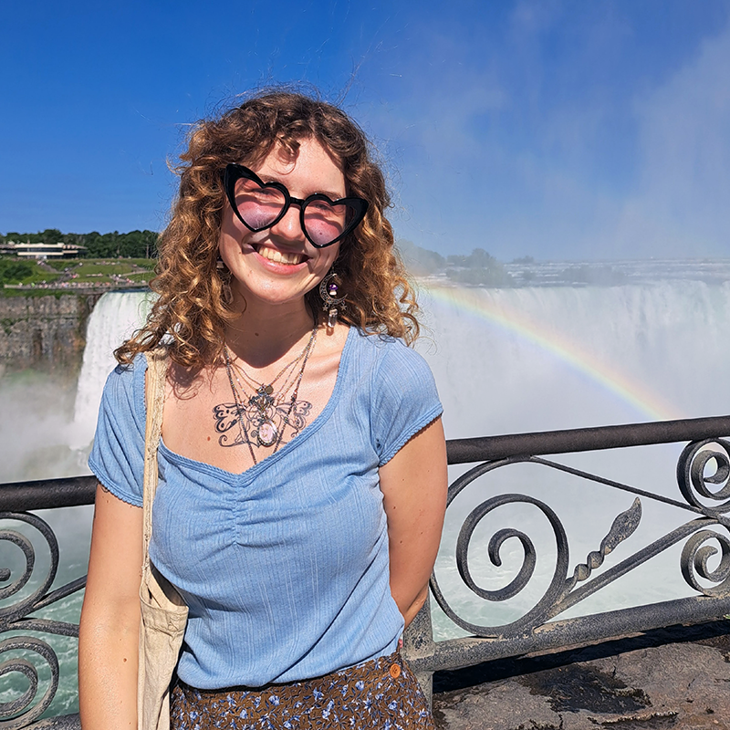 Isobel Flindall standing by a railing in front of Niagara Falls with a rainbow across the falls. They are wearing a blue shirt, necklace and heart-shaped sunglasses.
