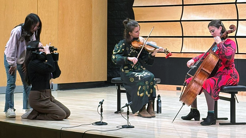 Two students on a stage filming two musicians on stringed instruments