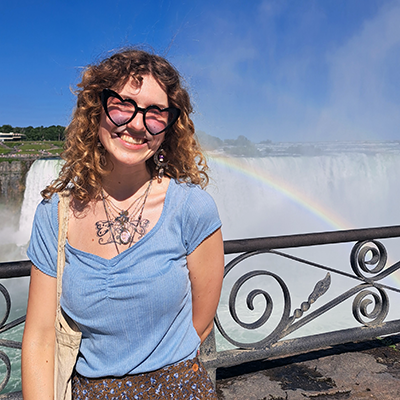 Isobel Flindall standing by a railing in front of Niagara Falls with a rainbow across the falls. They are wearing a blue shirt, necklace and heart-shaped sunglasses.
