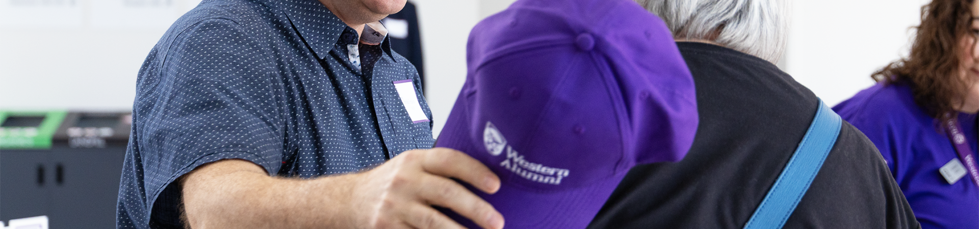 A person holding a Western Alumni hat. They are greeting another person warmly.