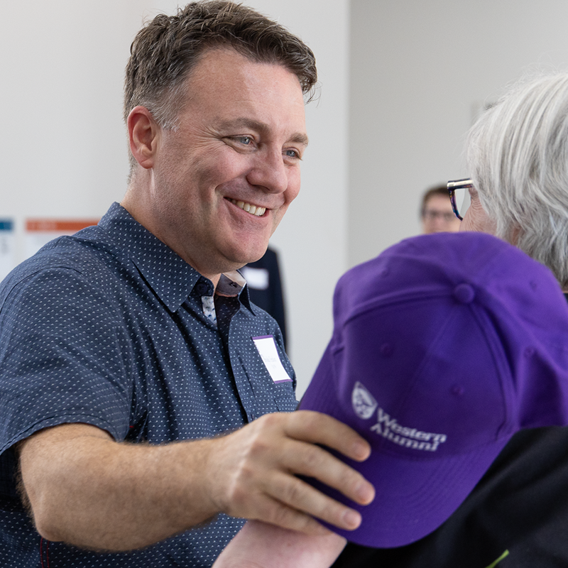 A person holding a Western Alumni hat. They are greeting another person warmly.
