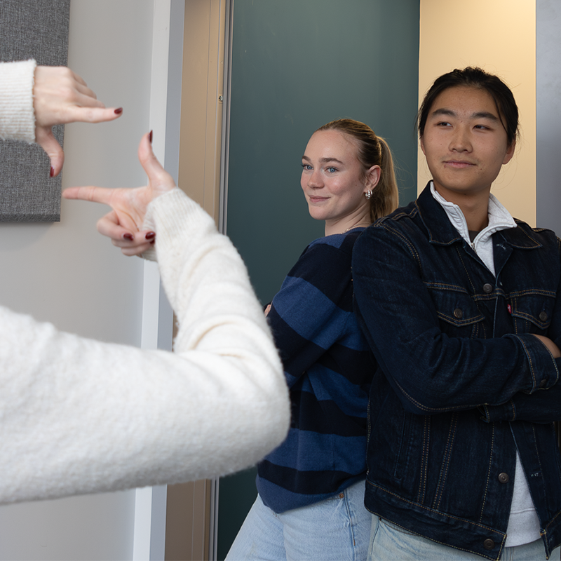 Two students standing back to back smiling at a person who is holding up their hands to frame the image